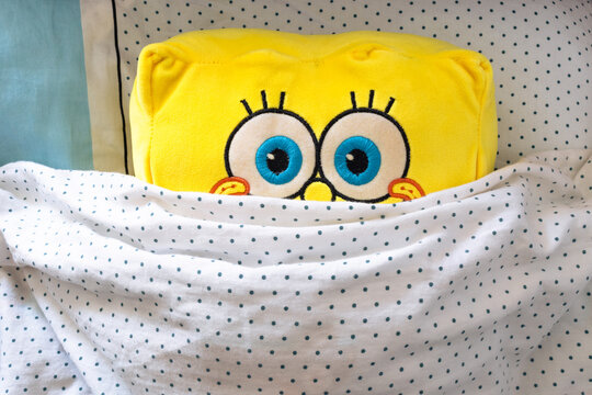 SpongeBob SquarePants peeks out from under the blanket. A plush toy SpongeBob lies in the bed under the blanket
