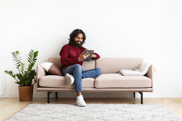 Cheerful young indian man sitting on couch, using digital tablet