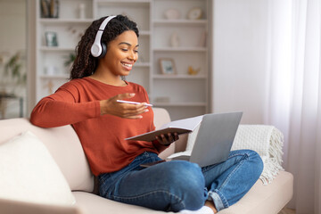Smiling black woman multitasking at home, writing in notebook while using laptop