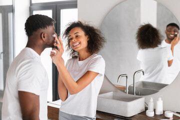 Young black couple shares flirty moment moisturizing skin in bathroom - obrazy, fototapety, plakaty