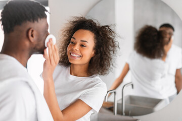 African wife applies skincare products to husband's face at bathroom