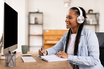 Smiling black female student with headphones taking notes from online lecture