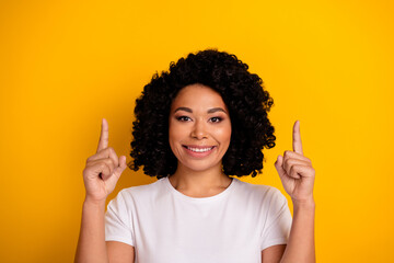 Portrait of cheerful girl with curly hairdo wear white t-shirt indicating up at sale empty space isolated on vivid yellow color background