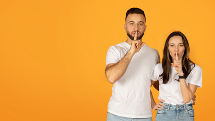 Quietly confident man and woman in white shirts placing fingers on lips in a 'shush' gesture