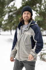 A man wearing a blue and gray jacket and a black hat is smiling. He is standing in the snow with trees in the background