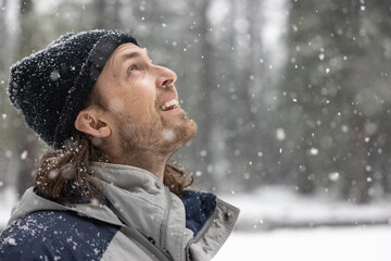 A man wearing a black hat and a gray jacket is smiling as he looks up at the snow falling from the sky