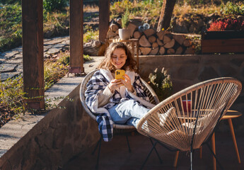 Young woman in a warm blanket on the terrace of a country house cottage enjoying the early spring sun, with smartphone - 751562777