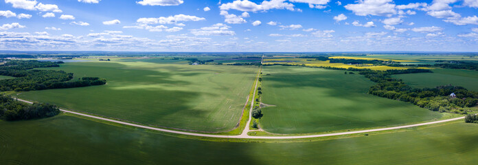 Aerial panoramic view of a vast prairie agricultural landscape with wheat fields. Gravel roads cross the image and the sky is pale blue with scattered white clouds.
