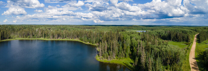 Aerial panoramic view of a beautiful northern park with a small lake, a forest of spruce trees, and a narrow gravel road. The sky is filled with puffy clouds.
