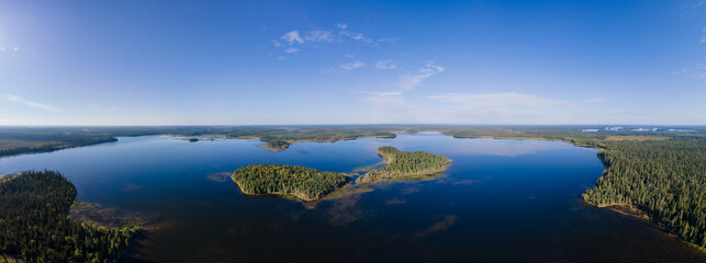 Aerial panoramic view of a northern wilderness lake with small islands. The water is calm and reflecting the blue sky.  The surrounding forest is a mix of spruce, pine, and aspen trees.
