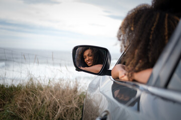 Portrait of a lovely young black woman through the rear view mirror as she looks out of her car window with the sea and sky in the background.