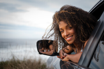 Portrait of a charming young black woman leaning out of the window of her car with the sea and sky in the background.