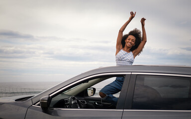 Blissful young black woman leaning out of the sunroof of her car with the sea and sky in the background. Female driver enjoying the freedom of a getaway. - 751568777