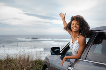 Blissful young black woman leaning out of the window of her car with the sea and sky in the background. Female driver enjoying the freedom of a getaway. - 751568798