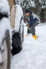 A man in a blue and gray jacket is shoveling snow from the ground. He is wearing yellow boots and a black hat