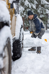 A man in a black hat and blue jacket is shoveling snow off the back of a truck