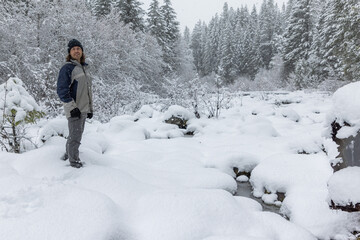 A man stands in the snow with his hands in his pockets. The snow is deep and the trees are bare. The man is wearing a blue jacket and a hat. The scene is quiet and peaceful