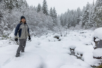 A man is standing in the snow with his hands in his pockets. He is wearing a blue jacket and a hat. The snow is deep and the trees are covered in snow