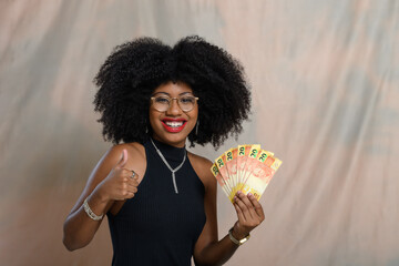 Black woman holding Brazilian money smiles, looks at the camera and makes a positive hand gesture.