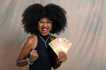 A black woman holds up Brazilian money, smiles, looks at the camera and celebrates with her hand.
