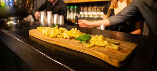 Sliced fresh lemons artistically presented on a wooden board in a bar setting, with bartenders in soft focus behind