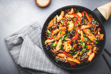 Asian stir fry chicken  slices with red paprika, mushrooms, chives and sesame seeds in frying pan. Gray kitchen table background, top view