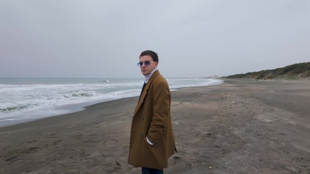 Charming well dressed Italian model actor ready for film scene during film production walks on the beach on a cloudy day and smiles looking at the camera.