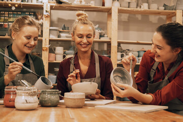 A company of three cheerful young women friends are painting ceramics in a pottery workshop.