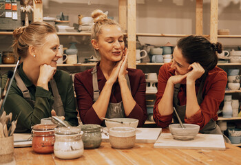 A company of three cheerful young women friends are painting ceramics in a pottery workshop.