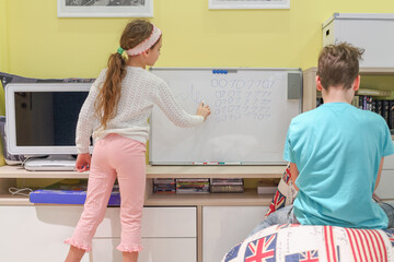Two teenagers in room, girl writes something on white board marker, seen from behind