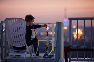 Young man adjusts burning of charcoal at hookah sitting in armchair on highrise roof at sunset dusk