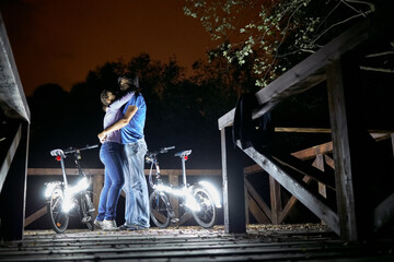 Man and  woman stand embracing at night on wooden platform near bicycles decorated with luminous diodes - Powered by Adobe
