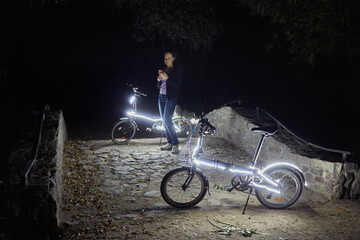 Woman with cell phone stands at night park near two bicycles decorated with luminous diodes