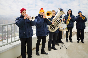 Brass band of six musicians play on roof of tall building at winter day