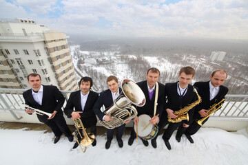 Brass band of six musicians in suits smile on roof of tall building at winter