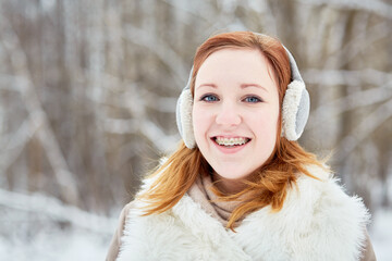 Humeral portrait of red-haired girl with braces on teeth in earmuffs at winter park