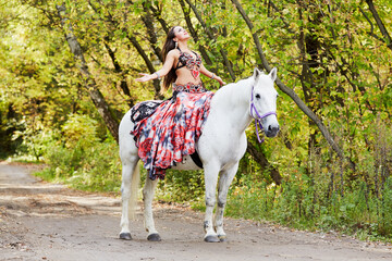 Young woman dressed in colourful dress decorated with playing cards sits on horseback with arms stretched to sides and face to sky in park