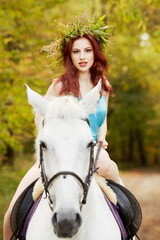 Red-haired woman with floral wreath on head rides on white horse in park