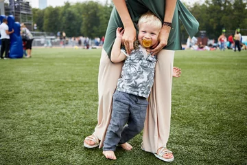 Poster Toddler boy with dummy in mouth stands barefoot with mother hands support on green grass © Pavel Losevsky