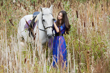 Girl in blue dress holds by bridle white horse among the reeds