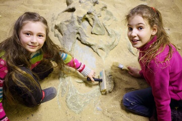 Two girls dig together in dry sand bones of dinosaur in sand