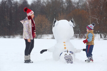Little boy and woman stand near upside down snowman at winter day
