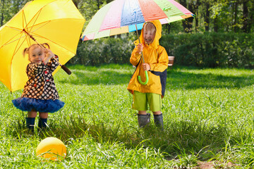 Brother and sister with colorful umbrellas in boots and jackets walk in park, ball are in grass