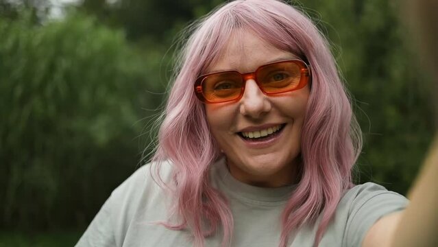 Caucasian joyful smiling beautiful young pink hair woman in orange sunglasses recording video for blog. Female talks on video call through webcam at backyard and waving hand 