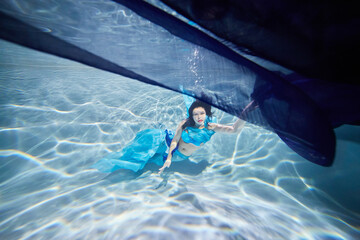 Young black-haired woman in blue dress poses underwater under dark-blue fabric with her eyes open - obrazy, fototapety, plakaty