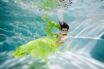 Smiling young woman in yellow-green dress swims posing in swimming pool underwater