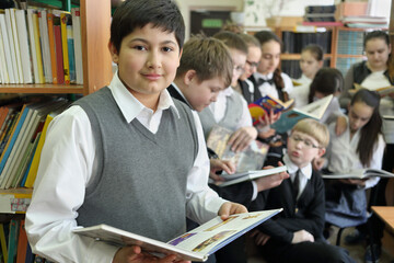 Ten students in school library reading books on background of shelves with books, one in foreground looking into camera, focus on boy left © Pavel Losevsky