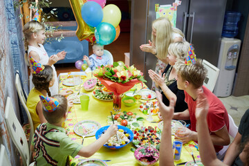 Seven adults and children sit at birthday table and congratulate little boy with applauses