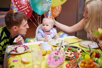 Little boy and parents in birthday caps sit at birthday table. Happy Birthday and Congratulations inscriptions on air-balloons.