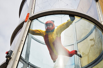Woman in helmet and red-yellow suit flies in air-tube with glass walls